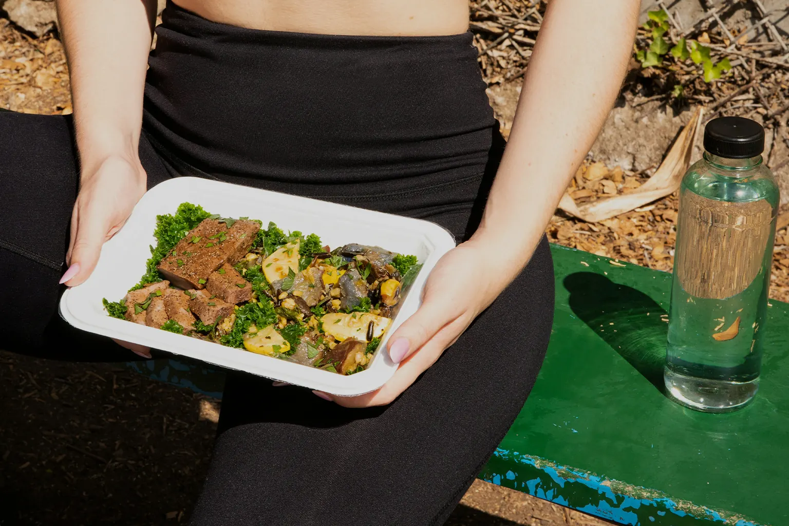 A tray of steak, kale, squash and mushrooms, held on a bench outdoors